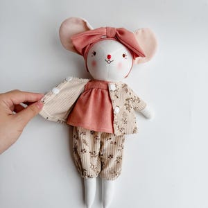 May include: A white fabric mouse doll wearing a pink headband, a pink dress, a beige cardigan with floral print, and beige pants with floral print.