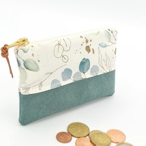 May include: A small, zippered pouch with a white fabric exterior featuring a floral pattern in shades of green and gold. The pouch has a teal faux leather bottom and a brown leather zipper pull.