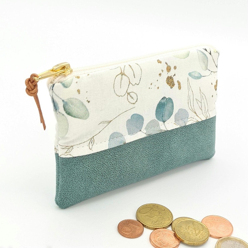 May include: A small zippered pouch with a white fabric exterior featuring a floral pattern in shades of green and gold. The pouch has a teal faux leather bottom.