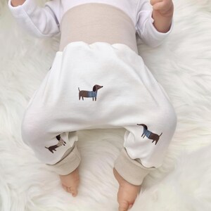 Könnte beinhalten: Weiße Babyhose mit braunen Dackel-Stickereien, die blau-weiß gestreifte Hemden tragen. Die Hose hat einen beigen Bund und Bündchen. Das Baby liegt auf einer weißen, flauschigen Oberfläche.