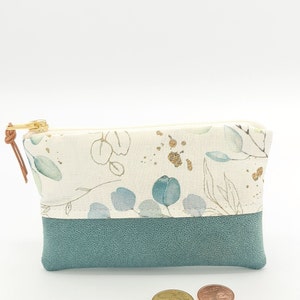 May include: A small, zippered pouch with a white fabric exterior featuring a floral pattern in shades of green and gold. The pouch has a teal blue faux leather bottom.