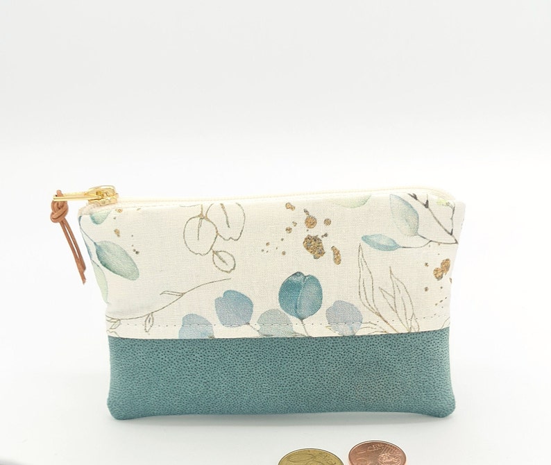 May include: A small, zippered pouch with a white fabric exterior featuring a floral pattern in shades of green and gold. The pouch has a teal blue faux leather bottom.
