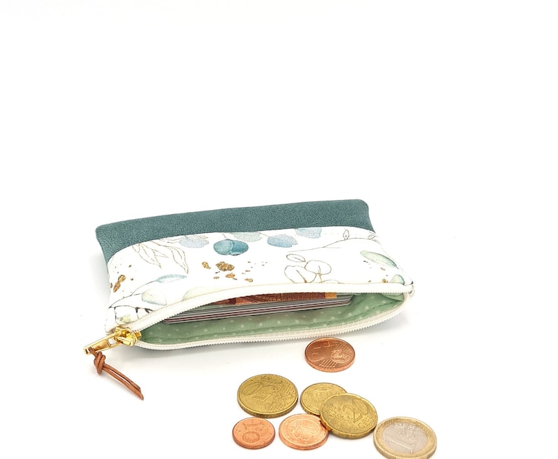 May include: A small teal zippered pouch with a white floral print and gold accents. The pouch is open and has a few euro coins spilling out.