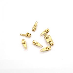 May include: A group of eight gold-colored metal zipper pulls scattered on a white surface.