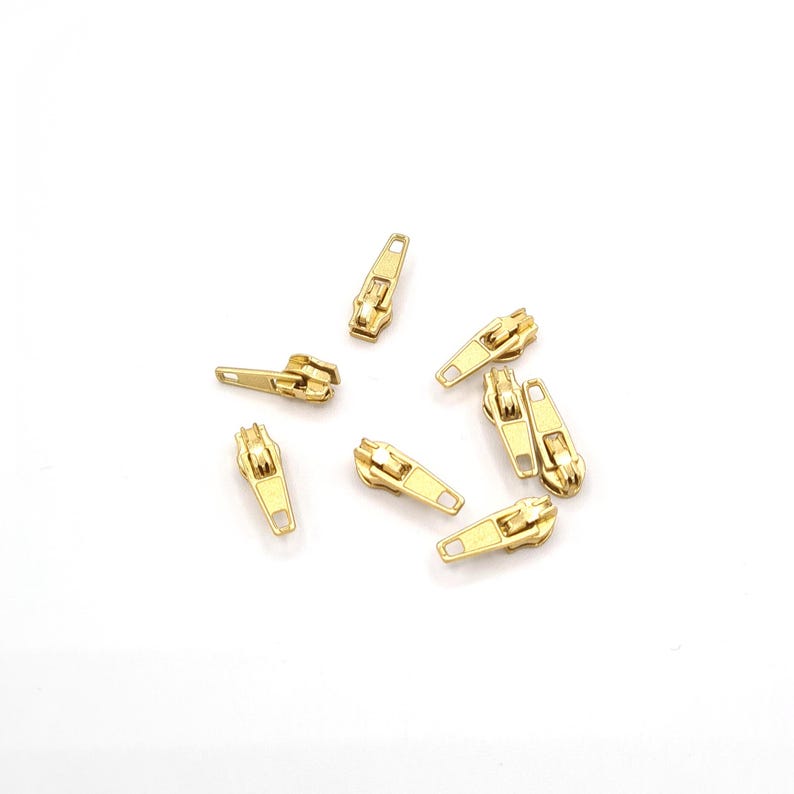 May include: A group of eight gold-colored metal zipper pulls scattered on a white surface.