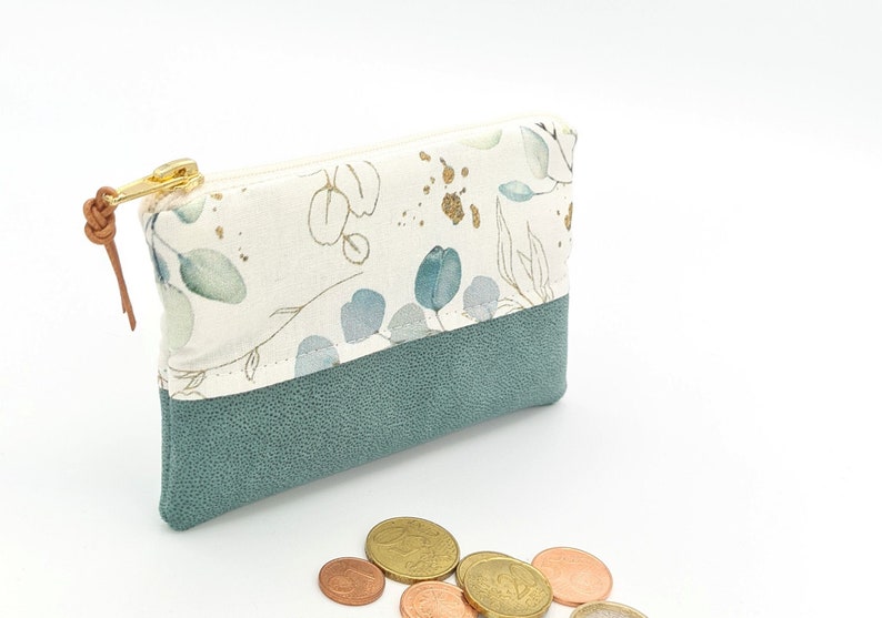 May include: A small, zippered pouch with a white fabric exterior featuring a floral pattern in shades of green and gold. The pouch has a teal faux leather bottom and a brown leather zipper pull.