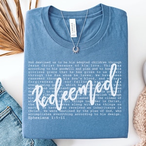 Christian women’s t shirt with the word Redeemed in white script lettering layered over Ephesians scripture text. Minimalist faith based tee with a soft, relaxed fit.