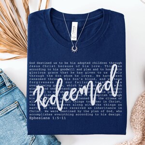 May include: Navy blue t-shirt with the word "Redeemed" in white script, along with a passage from Ephesians. A silver necklace with a sunburst pendant is on the shirt. The shirt is folded on a white surface with denim jeans and a beige sweater.