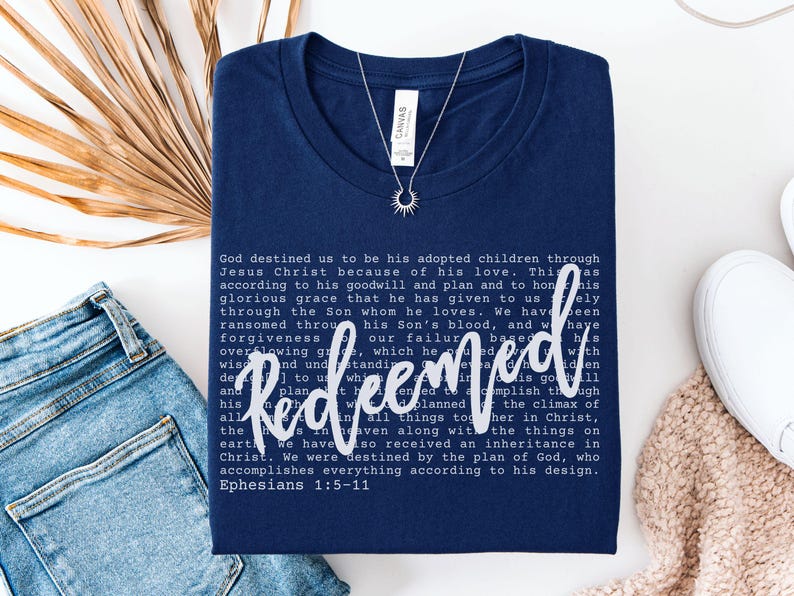 May include: Navy blue t-shirt with the word "Redeemed" in white script, along with a passage from Ephesians. A silver necklace with a sunburst pendant is on the shirt. The shirt is folded on a white surface with denim jeans and a beige sweater.
