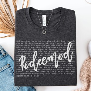 May include: Dark gray t-shirt with the word "Redeemed" in white script. The shirt features a printed passage from Ephesians 1:5-11. A silver necklace with a sunburst pendant is on the shirt.