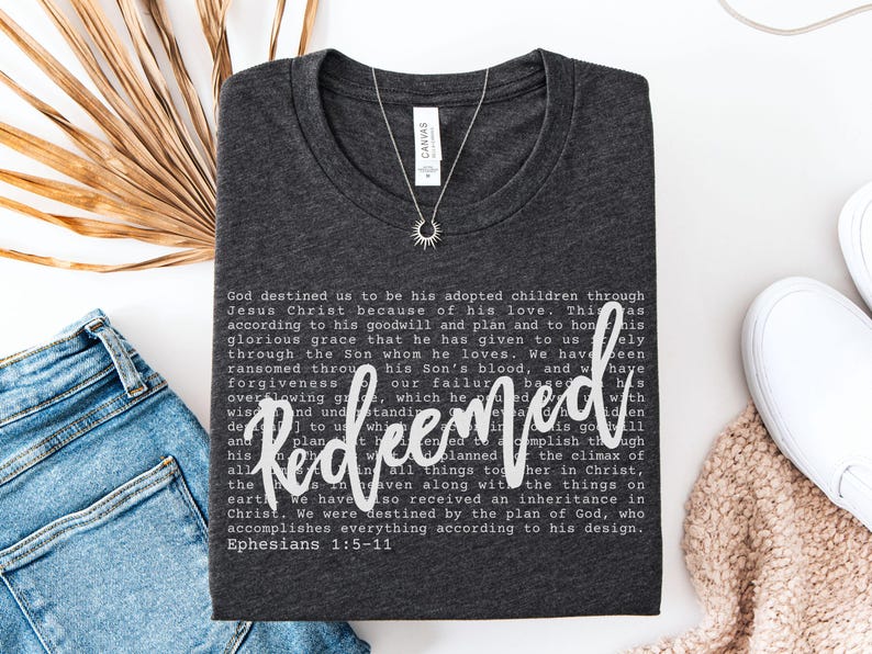 May include: Dark gray t-shirt with the word "Redeemed" in white script. The shirt features a printed passage from Ephesians 1:5-11. A silver necklace with a sunburst pendant is on the shirt.