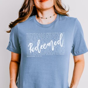May include: A light blue t-shirt with the word "Redeemed" in a cursive font. The shirt features a block of white text with a Bible verse. The shirt is a solid color and has short sleeves.