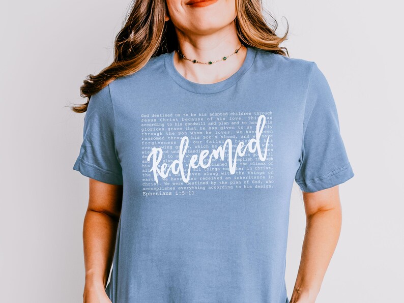 May include: A light blue t-shirt with the word "Redeemed" in a cursive font. The shirt features a block of white text with a Bible verse. The shirt is a solid color and has short sleeves.