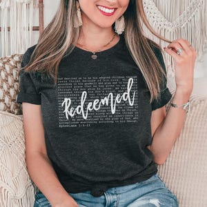 May include: Dark gray t-shirt with the word "Redeemed" in white script. The shirt also features a block of text in a smaller font. The shirt is worn by a person.