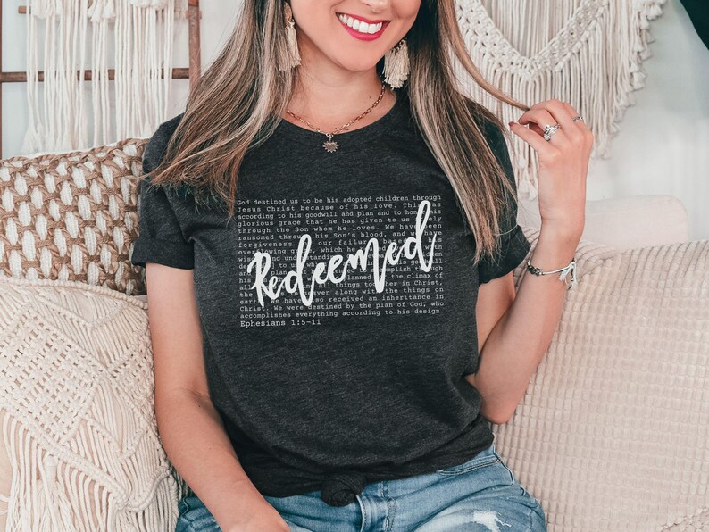 May include: Dark gray t-shirt with the word "Redeemed" in white script. The shirt also features a block of text in a smaller font. The shirt is worn by a person.