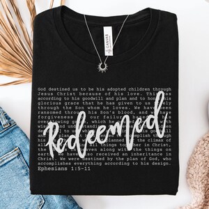 May include: Black t-shirt with the word "Redeemed" in white script. The shirt features a printed passage from Ephesians 1:5-11. A silver necklace with a sunburst pendant is visible.
