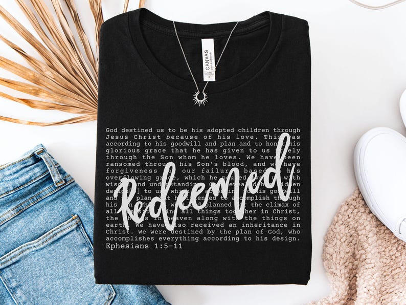 May include: Black t-shirt with the word "Redeemed" in white script. The shirt features a printed passage from Ephesians 1:5-11. A silver necklace with a sunburst pendant is visible.