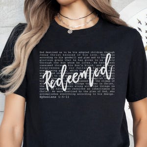 May include: Black t-shirt with the word "Redeemed" in white script. The shirt features a block of text in a smaller font, with the Bible verse Ephesians 1:5-11 at the bottom. The shirt is a casual wear item.