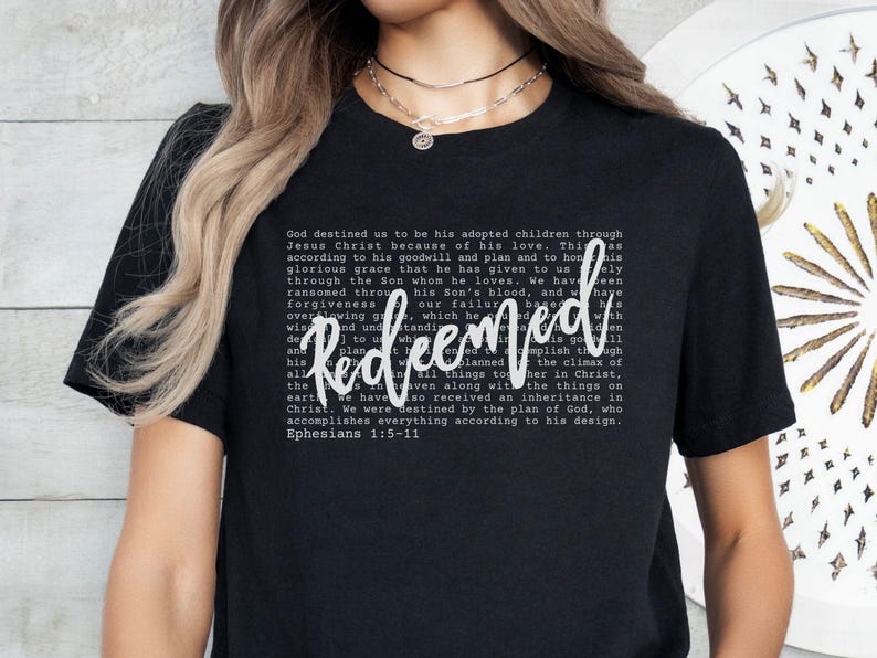 May include: Black t-shirt with the word "Redeemed" in white script. The shirt features a block of text in a smaller font, with the Bible verse Ephesians 1:5-11 at the bottom. The shirt is a casual wear item.
