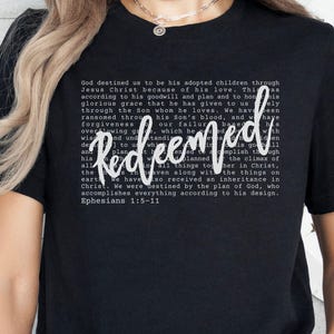 May include: Black t-shirt with the word "Redeemed" in a white script font. The shirt features a block of text in a smaller font, with the Bible verse Ephesians 1:5-11.