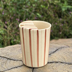 May include: A cylindrical ceramic tumbler with a cream-colored speckled base and vertical reddish-brown stripes. The cup features a matching ceramic lid that covers half of the top opening, including a small triangular cutout for sipping.