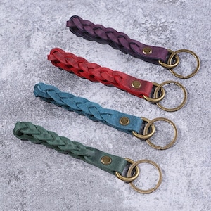Peut inclure: Quatre porte-clés en cuir tressé de couleurs violet, rouge, turquoise et vert. Chaque porte-clés est doté d'un anneau de couleur laiton et d'une lanière en cuir assortie fixée par un rivet de couleur laiton. Les porte-clés sont disposés en diagonale.