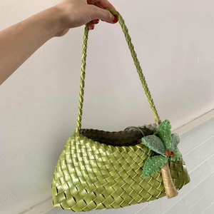 May include: A woven green handbag with a braided handle and a decorative felt palm tree. The bag has a rectangular shape and is held by a hand with a silver ring.