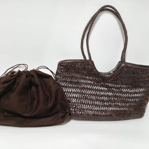 Puede incluir: Un bolso tote tejido marrón con asas trenzadas y una bolsa con cordón a juego. El bolso tiene un diseño único de tejido abierto, y la bolsa está hecha de una tela suave de color marrón oscuro. El bolso mide aproximadamente 38 cm de ancho.