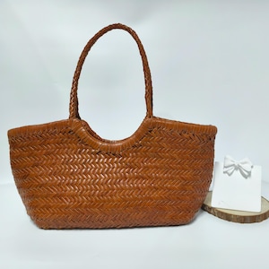 Può includere: Una borsa tote marrone intrecciata con un manico intrecciato. La borsa ha una forma rettangolare con un'apertura curva. Un fiocco decorativo bianco è su una piccola fetta di legno accanto alla borsa.