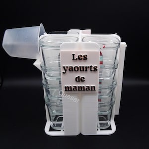 Peut inclure: Un support de pots de yaourt en plastique blanc avec quatre pots en verre. Le support porte l'inscription "Les yaourts de maman" et la marque "Quasar". Une tasse à mesurer en plastique transparent est attachée.