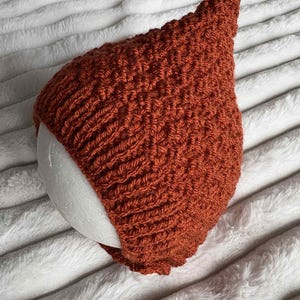 May include: A rust-coloured, hand-crocheted pixie hat. The hat has a pointed top and a textured stitch pattern. The hat is resting on a white, rounded form, set against a soft, white, textured background. The hat appears to be made of a soft yarn.
