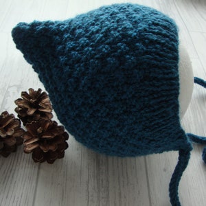May include: A teal blue knitted baby bonnet with ties. The bonnet is made with a textured stitch and has a pointed crown.