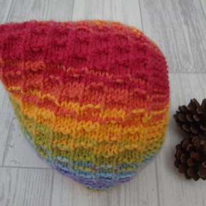 May include: A rainbow striped knitted hat with a pointed top. The hat is made with a textured stitch and features a variety of colours, including red, orange, yellow, green, blue, and purple.