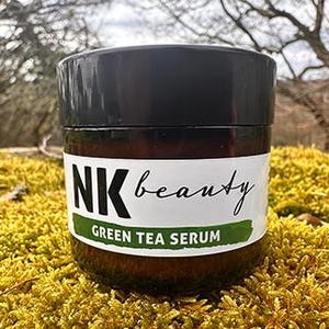 May include: A brown glass jar with a black lid, labelled "NK beauty Green Tea Serum." The jar is set on yellow-green moss, with a blurred natural background.