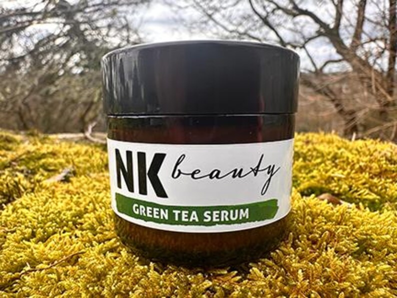 May include: A brown glass jar with a black lid, labelled "NK beauty Green Tea Serum." The jar is set on yellow-green moss, with a blurred natural background.