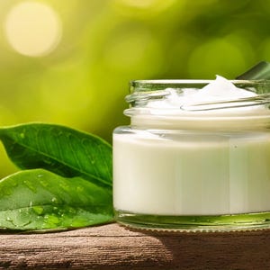 May include: A small glass jar containing white cream, with the lid ajar. The jar is on a wooden surface, beside green leaves. The background is a blurred green, creating a natural, organic feel.