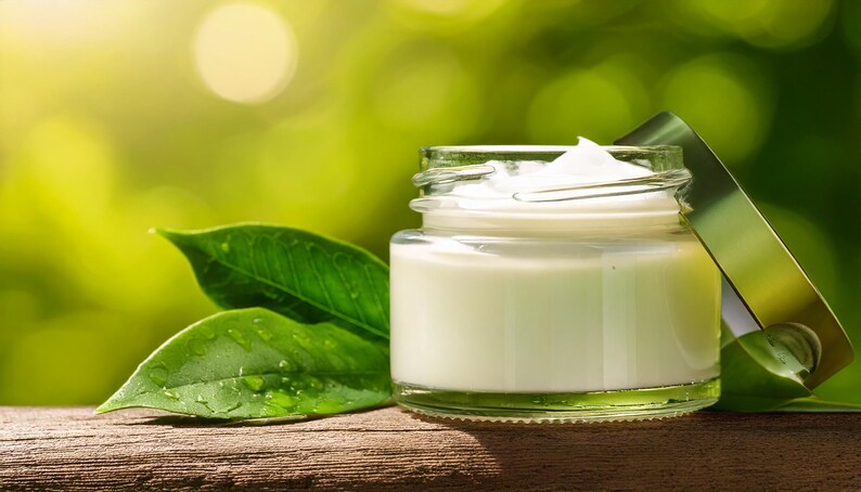 May include: A small glass jar containing white cream, with the lid ajar. The jar is on a wooden surface, beside green leaves. The background is a blurred green, creating a natural, organic feel.