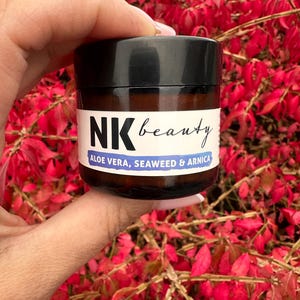 May include: A small, round jar of NK beauty product. The jar is dark brown with a black lid and a white label. The label reads "NK beauty" and lists ingredients: "ALOE VERA, SEAWEED & ARNICA". The background is filled with red leaves. The jar is held in a hand.