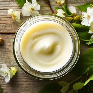 May include: A glass jar filled with cream, with a swirl pattern, alongside white flowers and green leaves. The cream is pale yellow, and the jar is transparent. The background is a wooden surface.