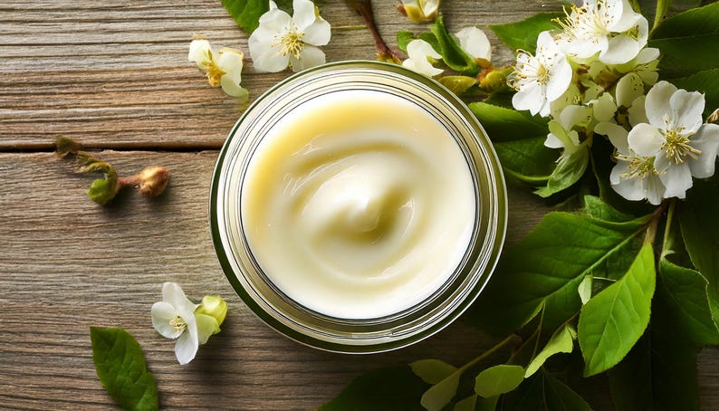 May include: A glass jar filled with cream, with a swirl pattern, alongside white flowers and green leaves. The cream is pale yellow, and the jar is transparent. The background is a wooden surface.