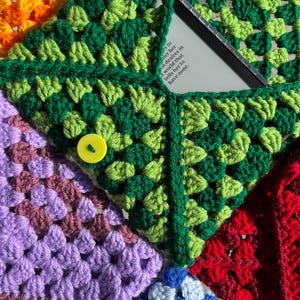 May include: A colorful, crocheted granny square bag with a green pocket holding a book. The bag features squares in orange, green, purple, red, and blue, with a yellow button closure. The book's text is partially visible.
