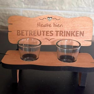 Könnte beinhalten: Ein Holz-Schnapsglashalter in Form einer Bank, mit zwei durchsichtigen Schnapsgläsern. Die Bank hat eine hellbraune Sitzfläche und Rückenlehne mit schwarzen Beinen. Auf der Rückseite der Bank steht der deutsche Text "Heute hier: BETREUTES TRINKEN".