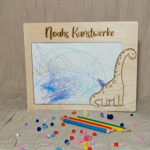 Könnte beinhalten: Ein Holzrahmen mit einem Dinosaurier-Ausschnitt und dem Text "Noah's Kunstwerke" oben. Der Rahmen hält ein weißes Papier mit bunten Kritzeleien darauf. Um den Rahmen herum liegen bunte Pompons und farbige Stifte.