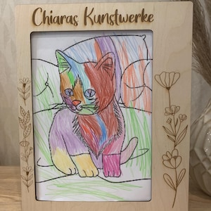 Könnte beinhalten: Ein Holzbildrahmen mit dem Text "Chiaras Kunstwerke" oben eingraviert. Der Rahmen enthält eine bunte Zeichnung einer Katze, die auf einem Kissen sitzt. Die Zeichnung ist von einem einfachen Blumendesign umgeben.