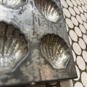 May include: Close-up of a vintage, rectangular metal madeleine pan. The pan has six shell-shaped molds with detailed ridges. The metal has a worn, aged appearance with areas of discoloration, suggesting its use over time. The pan is designed for baking madeleine cakes.