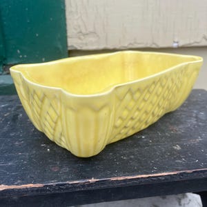 May include: A yellow ceramic planter with a woven pattern. The planter has a rectangular shape with rounded edges.