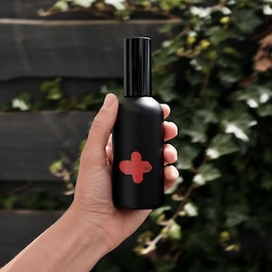 Könnte beinhalten: Eine schwarze Glasflasche mit schwarzem Verschluss und einem roten Pluszeichen auf der Vorderseite. Die Flasche wird in einer Hand gehalten.