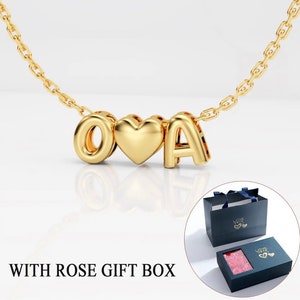Könnte beinhalten: Goldkette mit einem Herz und zwei Buchstaben, O und A, auf weißem Hintergrund. Die Kette wird in einer rosafarbenen Geschenkbox mit dem Wort "LOVE" geliefert.