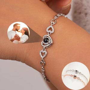 May include: A silver bracelet with a heart-shaped charm that projects a message when held up to a light. The charm is surrounded by smaller heart charms. The bracelet is shown on a person's wrist.