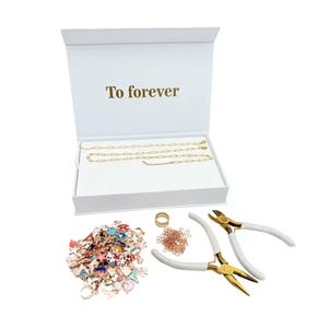 May include: A white jewellery-making kit with the text "To forever" on the inside lid. The kit includes gold-coloured chain necklaces, colourful charms, jump rings, a ring, and two white-handled pliers with gold-coloured heads.
